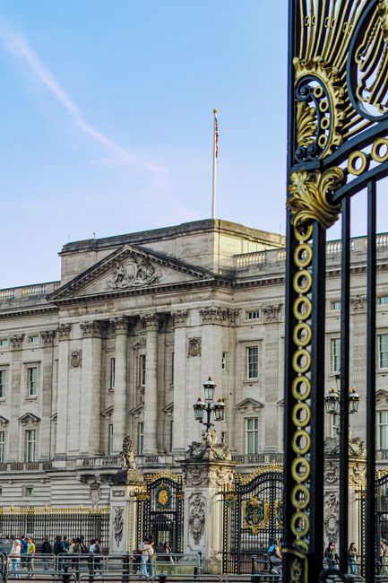 The image shows a historic stone building behind an ornate black and gold wrought iron gate, partially open, with the Union Jack flag flying on top of the building. In the foreground, there are severa