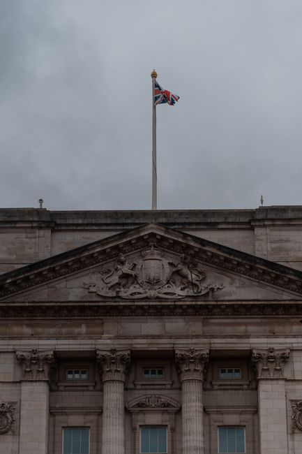 The image shows a historic stone building behind an ornate black and gold wrought iron gate, partially open, with the Union Jack flag flying on top of the building. In the foreground, there are severa