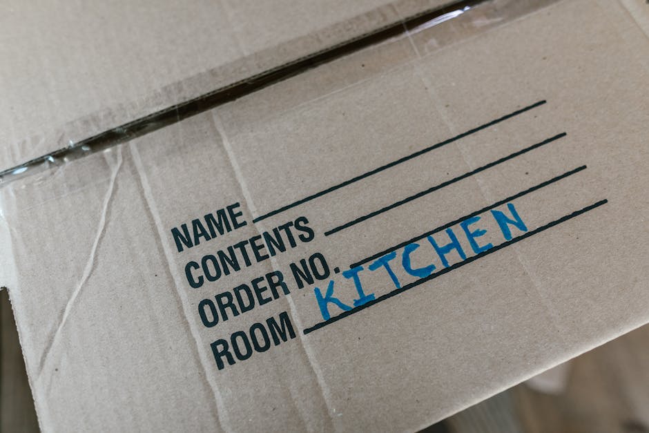 A person wearing a dark blue T-shirt and matching work trousers is seen handling a large cardboard box during a home relocation process inside an indoor setting, with natural light illuminating the sc